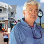 Man in a denim shirt with sunglasses stands on a seaside boardwalk, with a clock and beach ahead and a fries shop in the background.