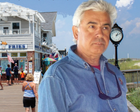 Man in a denim shirt with sunglasses stands on a seaside boardwalk, with a clock and beach ahead and a fries shop in the background.