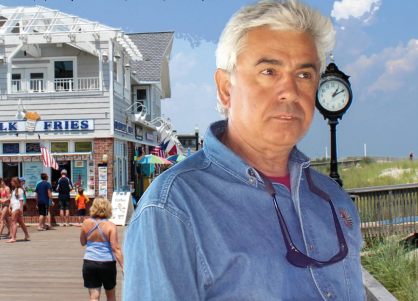 Man in a denim shirt with sunglasses stands on a seaside boardwalk, with a clock and beach ahead and a fries shop in the background.